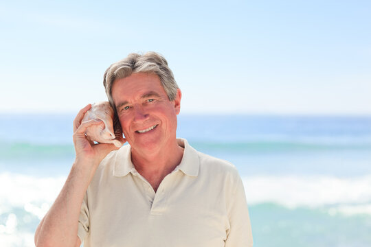 Senior man holding conch shell to right ear while standing at beach shoreline wearing light polo