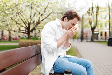 Fototapeta premium Man suffering from spring allergies, sneezing into his hands while sitting on a park bench surrounded by blooming trees. Seasonal hay fever concept.