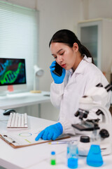 Female scientist wearing blue gloves and lab coat talking on cell phone while reviewing charts with...