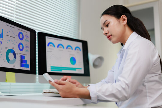 Young Asian businesswoman at her desk multitasking&mdash;analyzing financial charts and dashboards on dual monitors while using a smartphone for remote work and digital communication