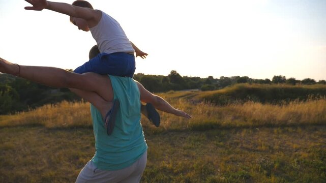 Little boy sitting on shoulders of his daddy and playing raised hands as airplane. Happy father and son playing on the field at sunset. Parent with kid having fun together outdoor. Slow motion