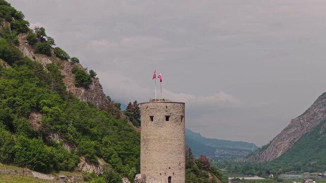 Cinematic drone footage of Ch&acirc;teau de la B&acirc;tiaz in Martigny, Switzerland, featuring the historic medieval tower, surrounding mountains and panoramic views over the alpine town on a sunny bluebird day.