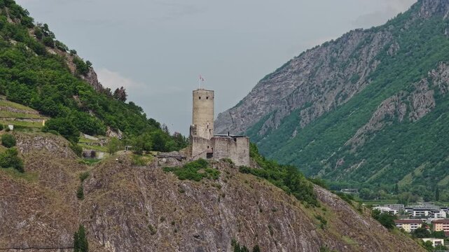 Cinematic drone footage of Ch&acirc;teau de la B&acirc;tiaz in Martigny, Switzerland, featuring the historic medieval tower, surrounding mountains and panoramic views over the alpine town on a sunny bluebird day.