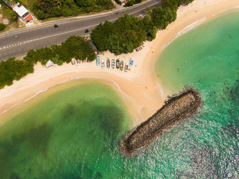 Aerial view of Devil's Rock and a sandy beach with colorful fishing boats, turquoise ocean water, a coastal road, and lush tropical trees Ahangama, Southern Province, Sri Lanka.