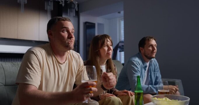 Three friends sitting together in a cozy living room watching an intense sports game on TV, enjoying drinks and snacks while reacting excitedly and celebrating moments