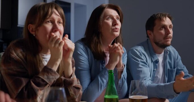 Three adults sitting closely together at a table indoors, watching something with focused expressions that turn into smiles and laughter, holding drinks and enjoying a social moment