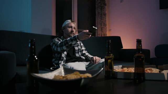 Young man in plaid shirt and cap pointing TV remote while relaxing on floor against couch. Beer bottles, chips bowl and pizza box in foreground. Moody blue warm light. Channel surfing 