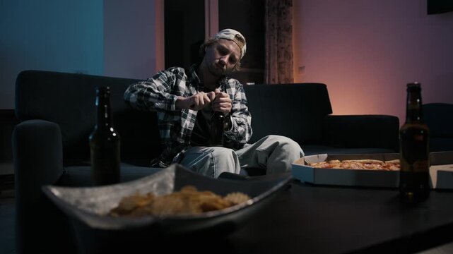Young man in plaid shirt and cap sitting against couch opening beer bottle. Chips bowl, pizza box and beer bottles on coffee table in foreground. Moody blue warm TV light. Solo sports watching night 