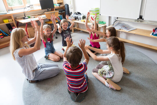 Teacher and pupils sitting in a circle on floor of their classroom raising hands