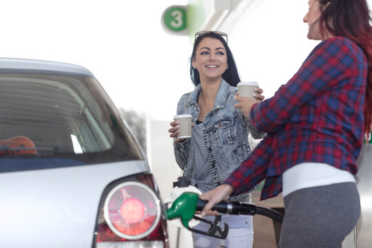 Two women with takeaway coffee fueling car at gas station