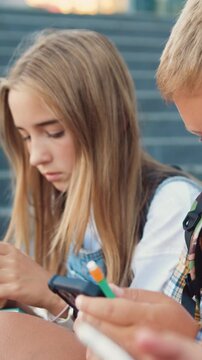 Close up of group of schoolchildren sitting on stairs outdoors using mobile phone communicating online. Diverse kids use internet on cellphone while relaxing on street at sunny day. Slow motion