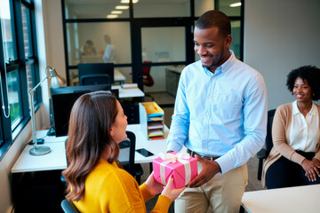 Naklejka premium African American young adult businessman giving gift box to Caucasian female coworker in office, celebrating workplace appreciation. Ideal for employee recognition, team culture marketing