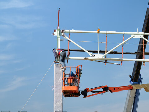 Construction workers wearing safety harnesses assemble metal roof framework from cherry picker. Industrial construction scene highlights engineering precision, workplace safety, teamwork and progress.