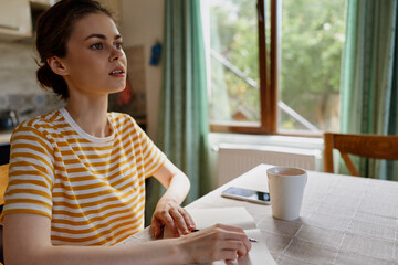 Woman with short hair wearing a striped shirt sits at a table near a window, writing in a notebook beside a mug. Daylight fills a calm, cozy home scene with relaxed focus.