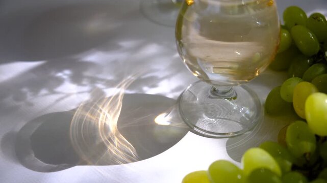 A chilled glass of dessert white wine reveals a pale golden colour, radiant in the morning light. Its aroma hints at notes of ripe peach and honey, echoing the fresh bunch of white grapes in sunlight