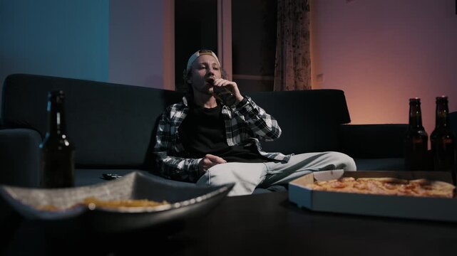 Young man jumps over couch and lands seated drinking beer while watching TV. Chips and pizza on table. Moody blue light. Playful energetic game night at home.
