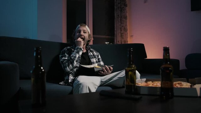Young man in plaid shirt eating chips on couch, staring at TV. Beer bottles and pizza box in foreground. Moody blue light. Solo sports fan snacking during match at home.
