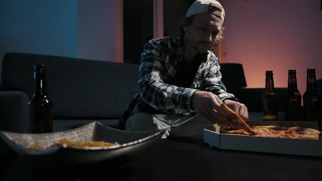 Young man takes pizza slice from box, eats and drinks beer cross-legged on couch. Chips, beer bottles, remote on table. Moody TV light. Relaxed solo sports night ritual at home.
