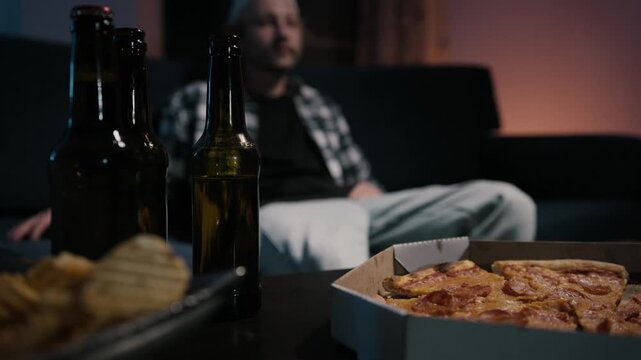 Sharp foreground of beer bottles, pizza box and chips bowl with blurred man settling on couch pointing TV remote behind. Moody game night atmosphere at home.
