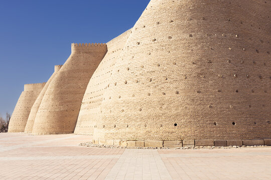 Ark Fortress walls in Bukhara with massive curved brick bastions under clear blue sky. Ideal for travel guides, history publications, and tourism marketing about Uzbekistan and the Silk Road.