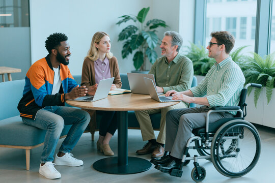 Wide shot of diverse business team collaborating with laptops during office meeting, including man with disability in wheelchair. Ideal for workplace inclusion, teamwork campaigns
