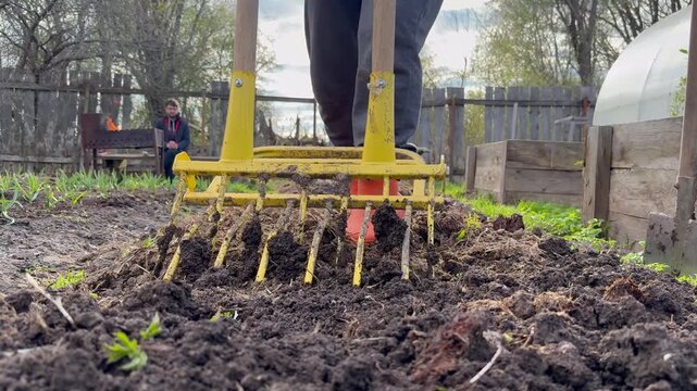 A close-up of a gardener in orange rubber boots using a manual garden fork (broadfork) or soil ripper to till the earth. 