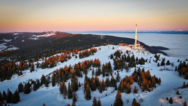 Aerial view of Col de la Faucille mountain pass featuring a communication tower, snow-covered slopes, and pine trees under a golden sunset sky Gex, Auvergne-Rhone-Alpes, France.