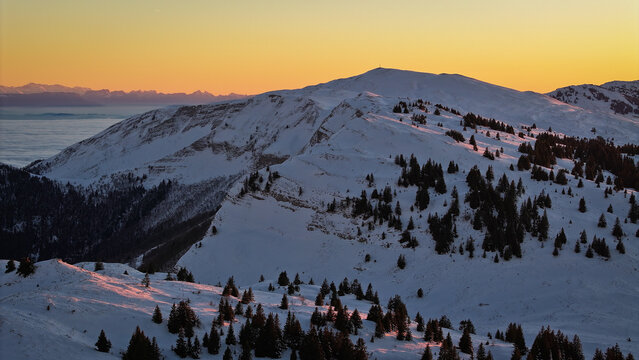 Aerial view of Col de la Faucille mountain pass with snow-covered slopes and pine trees under a golden sunset sky Gex, Auvergne-Rhone-Alpes, France.