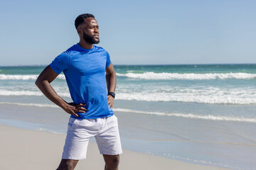 Fototapeta premium Mid-adult African American man standing on wet sand gazing toward sea in blue shirt, smartwatch