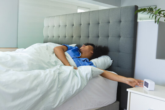 Middle-aged African American woman lying in bed wearing blue pajama top reaching for alarm clock