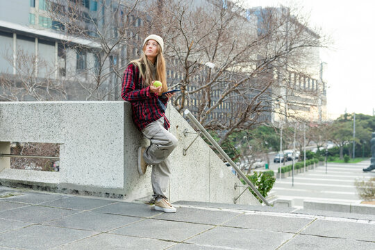 Woman leaning on parapet in urban plaza, wearing plaid jacket, with green apple and smartphone