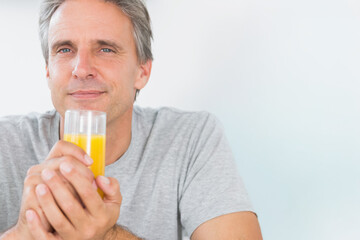 Fototapeta premium Middle-aged man sitting against pale wall holding clear glass of orange juice, wearing gray t-shirt