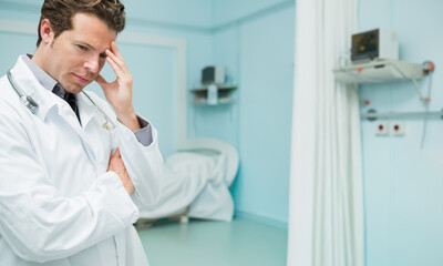 Fototapeta premium Male doctor wearing white coat, stethoscope, standing left, holding temple, viewing bed, copy space
