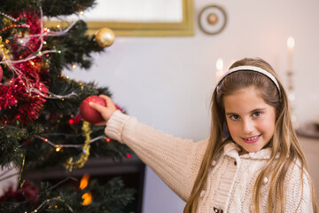 Fototapeta premium Placing large red bauble on Christmas tree in living room, child girl wearing knit sweater headband