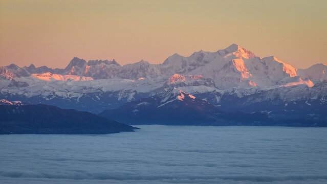 Aerial view of the snow-covered peaks of the Mont Blanc massif glowing in the golden light of sunset above a thick layer of clouds, Chamonix, Auvergne-Rhone-Alpes, France.
