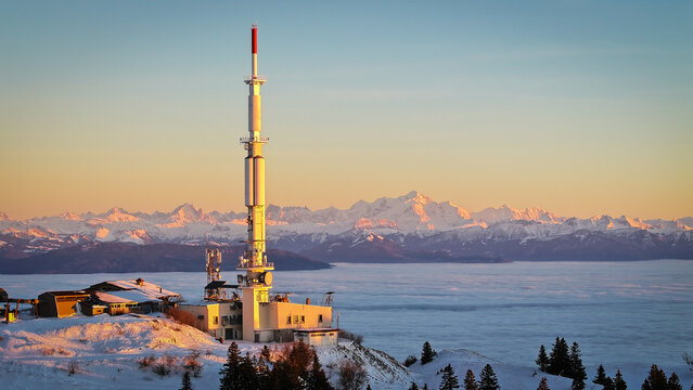 Aerial view of the Mont Blanc massif and a telecommunications tower on a snowy mountain ridge above a sea of clouds during sunset in Chamonix, Auvergne-Rhone-Alpes, France.