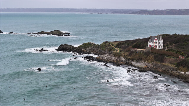 Aerial view of the rugged coastline of Locquirec featuring a white house with red shutters on a cliff overlooking the turquoise sea and surfers in the waves Locquirec, Brittany, France.