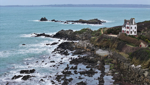 Aerial view of the rugged coastline of Locquirec featuring a white house with red shutters on a cliff overlooking turquoise waters and crashing waves Locquirec, Brittany, France.