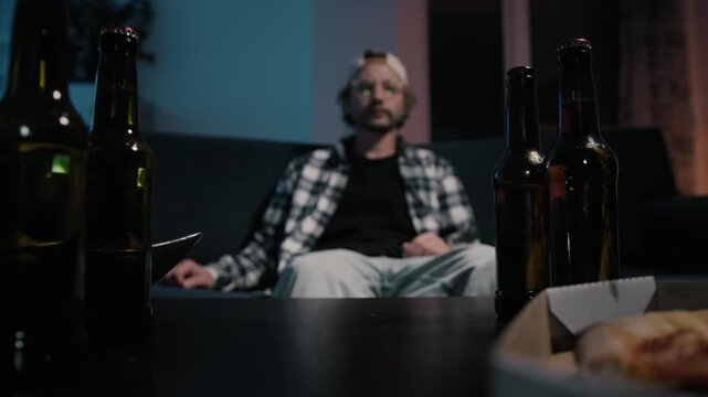 Rack focus shot with beer bottles and pizza in foreground while man sits on couch, grabs beer and drinks during late night television watching in dark cinematic apartment interior.