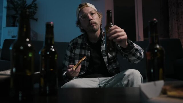 Medium shot of man eating pizza and drinking beer while watching television at night. Beer bottles in foreground and cinematic apartment lighting create relaxed home mood.