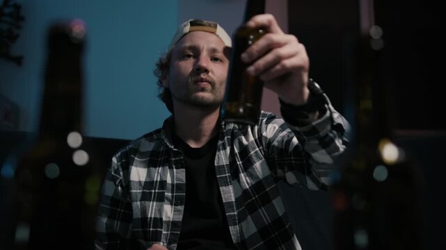 Medium close-up of man drinking beer while watching television at night, sitting on sofa in dark apartment with cinematic blue light and blurred bottles foreground.