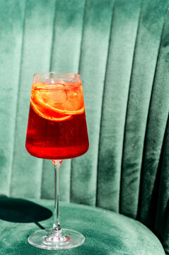 An orange cocktail with orange slices and ice in a stemmed glass sits on a green velvet channel-tufted armchair.