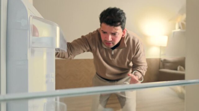 POV from inside fridge of young man opening refrigerator door with empty glass feeling disappointed looking for cold water or drink 