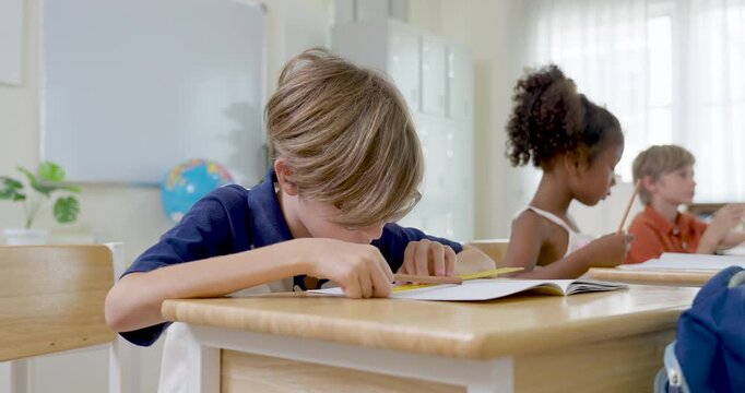 Slow motion view of young elementary pupils writing in notebook with pencil at wooden desk, diverse boy and girl doing classwork during back to school routine in bright modern primary classroom.