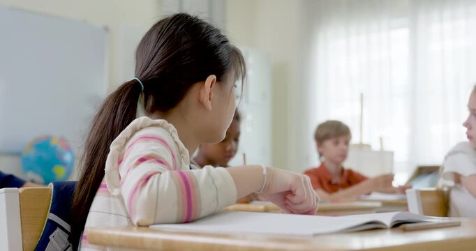 Asian elementary girl writes classwork in notebook while classmates study behind, first day classroom routine builds learning habit, concentration, school confidence, independent daily progress