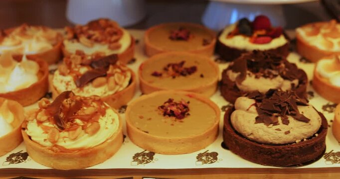 Assorted mini cakes put on tray in cafe dessert counter display. Sweet selection shows nut topping fruit slices with chocolate curls and soft cream swirls