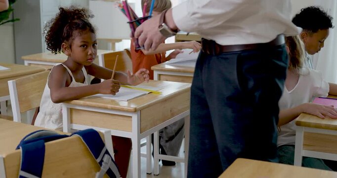 Adult male primary teacher walks between wooden desks during morning school routine, guiding multicultural children writing in notebooks, supporting discipline, classwork, study habit, order