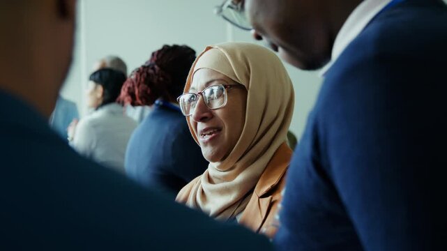 An enthusiastic woman in a hijab smiles and laughs, then listens intently among a diverse group of attendees at a business conference, showcasing engagement and active participation.