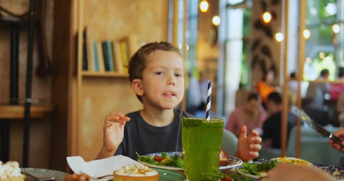 Little boy sits at cafe table pointing toward dessert with drink. Preschooler son asks question about food reacting playfully to family conversation