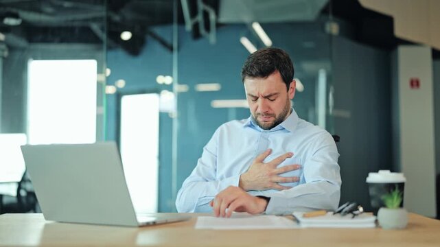 Unwell male entrepreneur holding hand on chest, suffering from sudden heart ache or severe panic attack while working at office laptop. Overworked corporate manager feeling sharp physical pain.